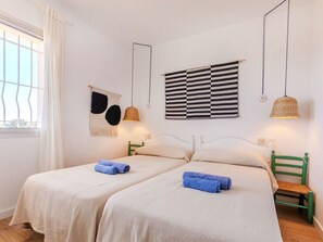 Habitació doble amb dos llits, una finestra amb cortines, un tapís negre i blanc a la paret i una làmpada penjant teixida.