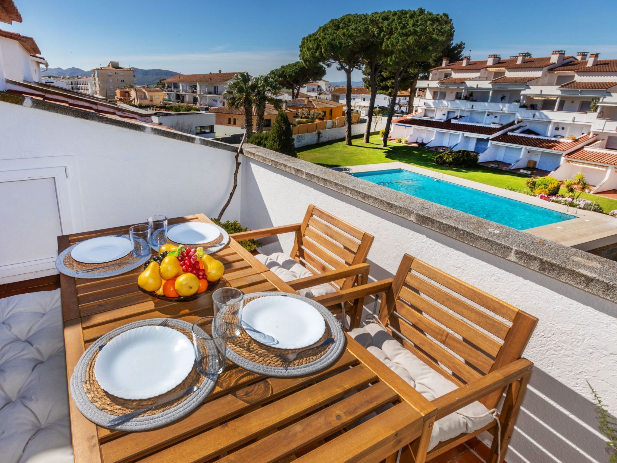 An outdoor dining area with a pool view, wooden furniture, and a table set with plates and fruit.