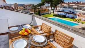 An outdoor dining area with a pool view, wooden furniture, and a table set with plates and fruit. - Residencial Gran Sol by Interhome (L'Escala)