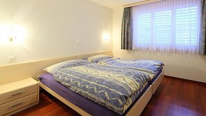 Een slaapkamer met een bed, nachtkastje, lade, raam met gordijnen en een plafondlamp.