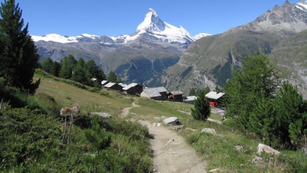 Un sentier de montagne qui mène à un groupe de maisons traditionnelles en bois, avec des sommets enneigés en arrière-plan.