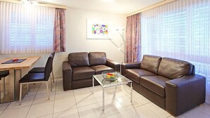 Een moderne woonkamer met een bruine leren bank, een glazen salontafel, een eettafel met stoelen en grote ramen voorzien van gordijnen.