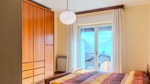 A bedroom with a wooden wardrobe, a bed with a colorful quilt, a wooden table, and a view of a balcony with a railing. - Apartment Marta by Interhome (Cerano d'Intelvi)
