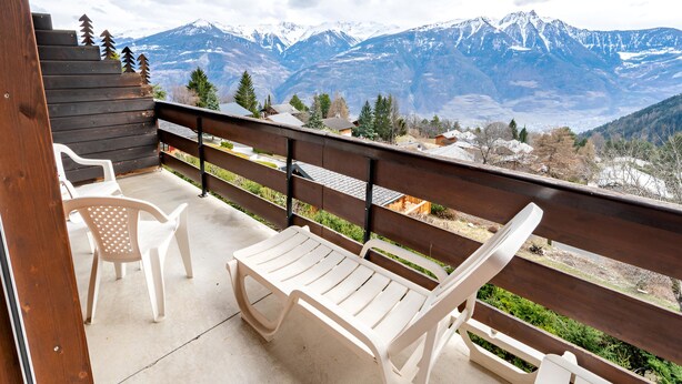 Un balcon avec deux chaises en plastique blanc et une table en bois, d’où l’on peut admirer un paysage montagneux avec des sommets enneigés et un ciel dégagé.