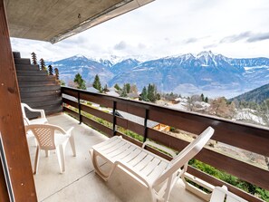 A balcony with two white plastic chairs and a wooden table, overlooking a mountainous landscape with snow-capped peaks and a clear sky.