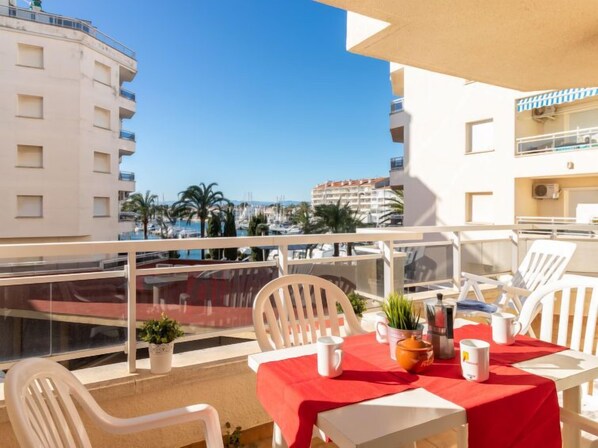Ein Balkon mit weißen Stühlen und einem Tisch, der mit einer roten Tischdecke gedeckt ist, mit Blick auf einen Yachthafen mit Booten und Palmen.