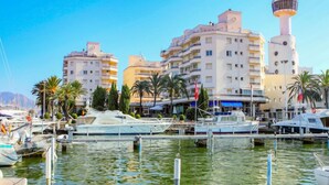 A marina with boats docked, surrounded by multi-story buildings with balconies, palm trees, and a prominent tower structure. - Club Nautic by Interhome (Empuriabrava)