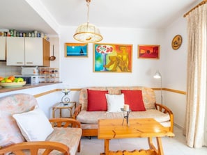 Ein gemütliches Wohnzimmer mit einer Sofagarnitur, einem Holztisch und einem Esstisch. An den Wänden hängen drei Bilder, eine Deckenleuchte und ein Küchenbereich mit Schränken und einer Obstschale.