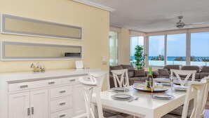 Uma sala de jantar moderna com uma mesa branca posta para quatro pessoas, um armário branco com gavetas e uma janela grande com vista para o oceano.