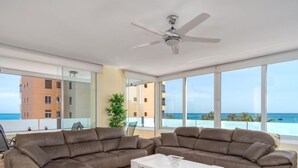 Uma sala de estar moderna com sofá seccional, mesa de centro branca e ventilador de teto. Grandes janelas proporcionam vista para o oceano e edifícios.