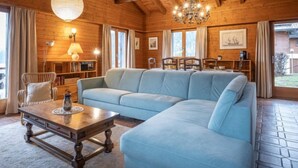Un acogedor interior de cabaña de madera con un amplio sofá seccional azul claro, vigas de madera en el techo, una araña y un comedor con mesa y sillas.
