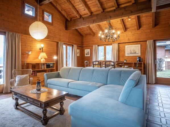 A cozy log cabin interior with a large light blue sectional sofa, wooden ceiling beams, a chandelier, and a dining area with a table and chairs.