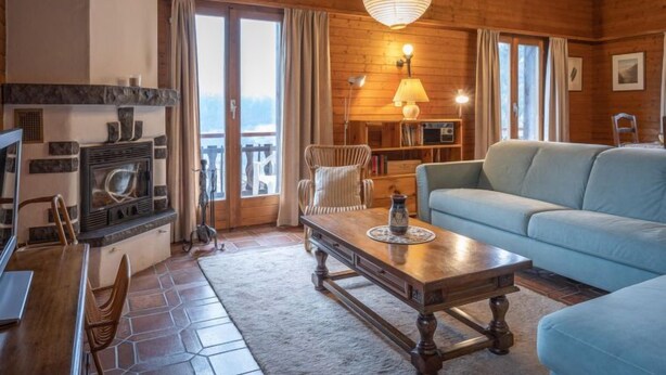 Interior acogedor de una cabaña de madera con chimenea de piedra, un sofá, una mesa de comedor y un ventanal con vistas panorámicas.