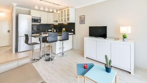 A modern kitchen with white cabinets, a black countertop, and a built-in microwave. There is a dining area with a table and chairs, a TV on a white cabinet, and a small plant on the table. - Le Beach Garden by Interhome (Cannes)
