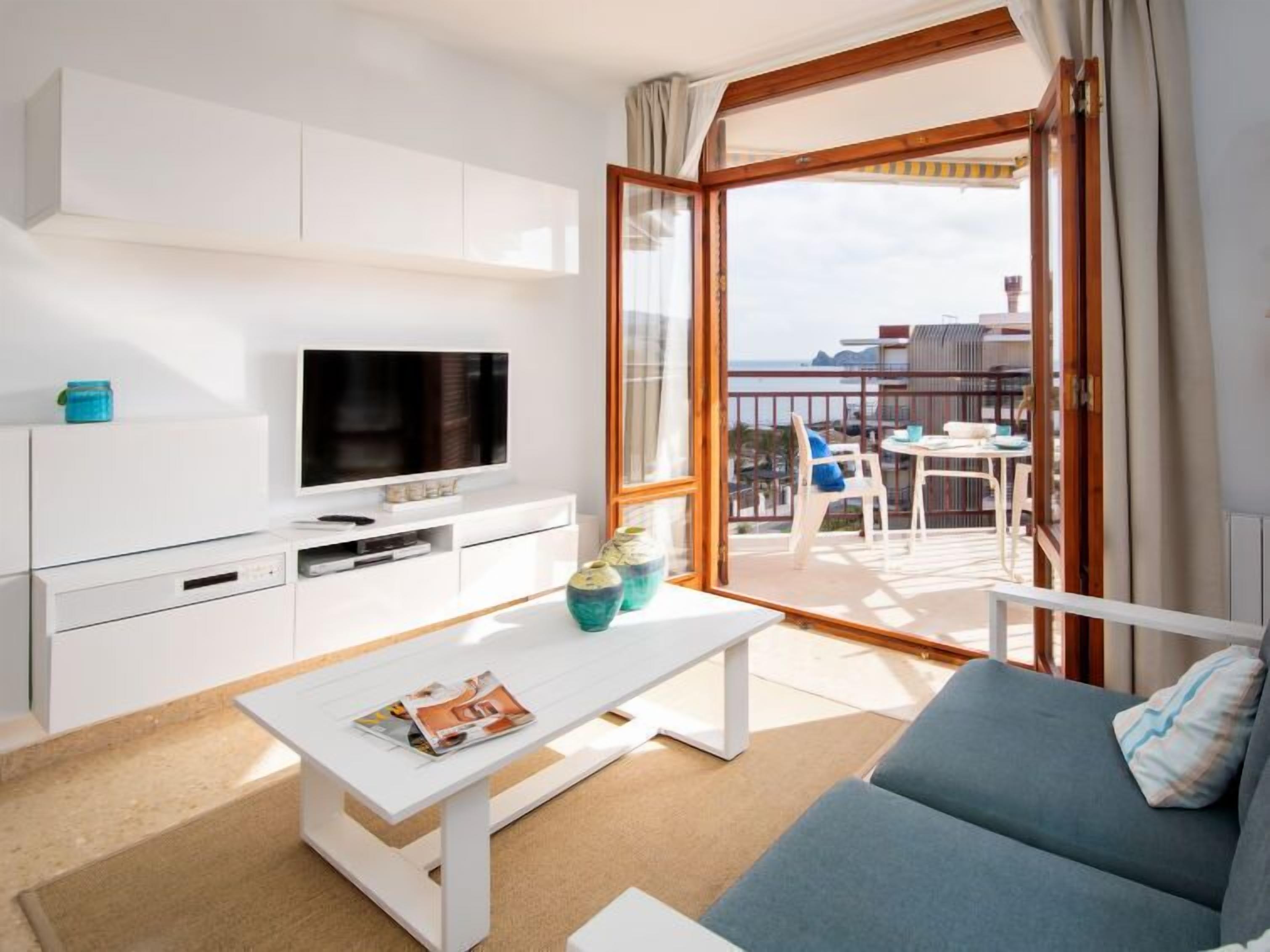 A modern living room with a flat-screen TV, white furniture, a balcony with a table and chairs, and a view of the city.