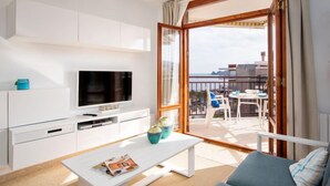 Una sala de estar moderna con televisor de pantalla plana, muebles blancos, un balcón con mesa y sillas, y vista a la ciudad.