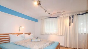 Un dormitorio con cama de madera, colchón azul, cortinas blancas y un ventilador de techo.