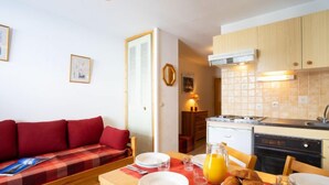 A cozy living area with a red sofa, a wooden dining table set with plates, glasses, and a jug of orange juice. A kitchen with wooden cabinets and a stove is visible in the background. - Plein Soleil (Val Claret) by Interhome (Tignes)