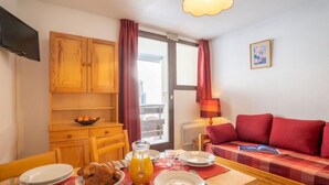 A cozy living room with a red sofa, wooden cabinets, a dining table set for breakfast, and a window with curtains. - Plein Soleil (Val Claret) by Interhome (Tignes)