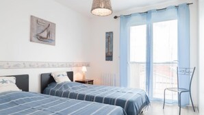 A bedroom with two beds, a window with light blue curtains, a chair, and a hanging light fixture. - Patguyflor by Interhome (Mimizan)