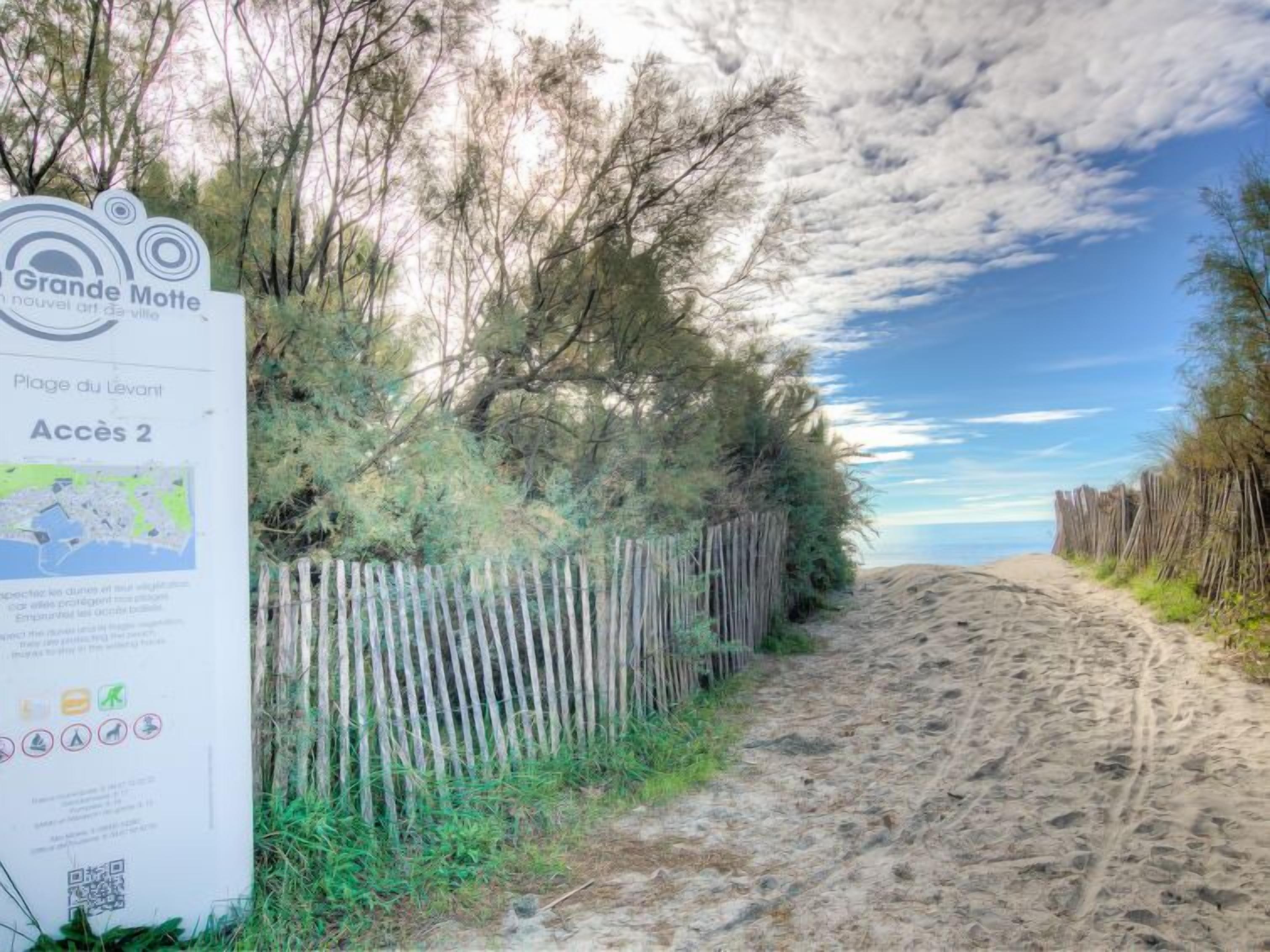 Un sentier bordant la plage, avec une clôture en bois et un panneau indiquant « Grande Motte » et « Accès 2 ».