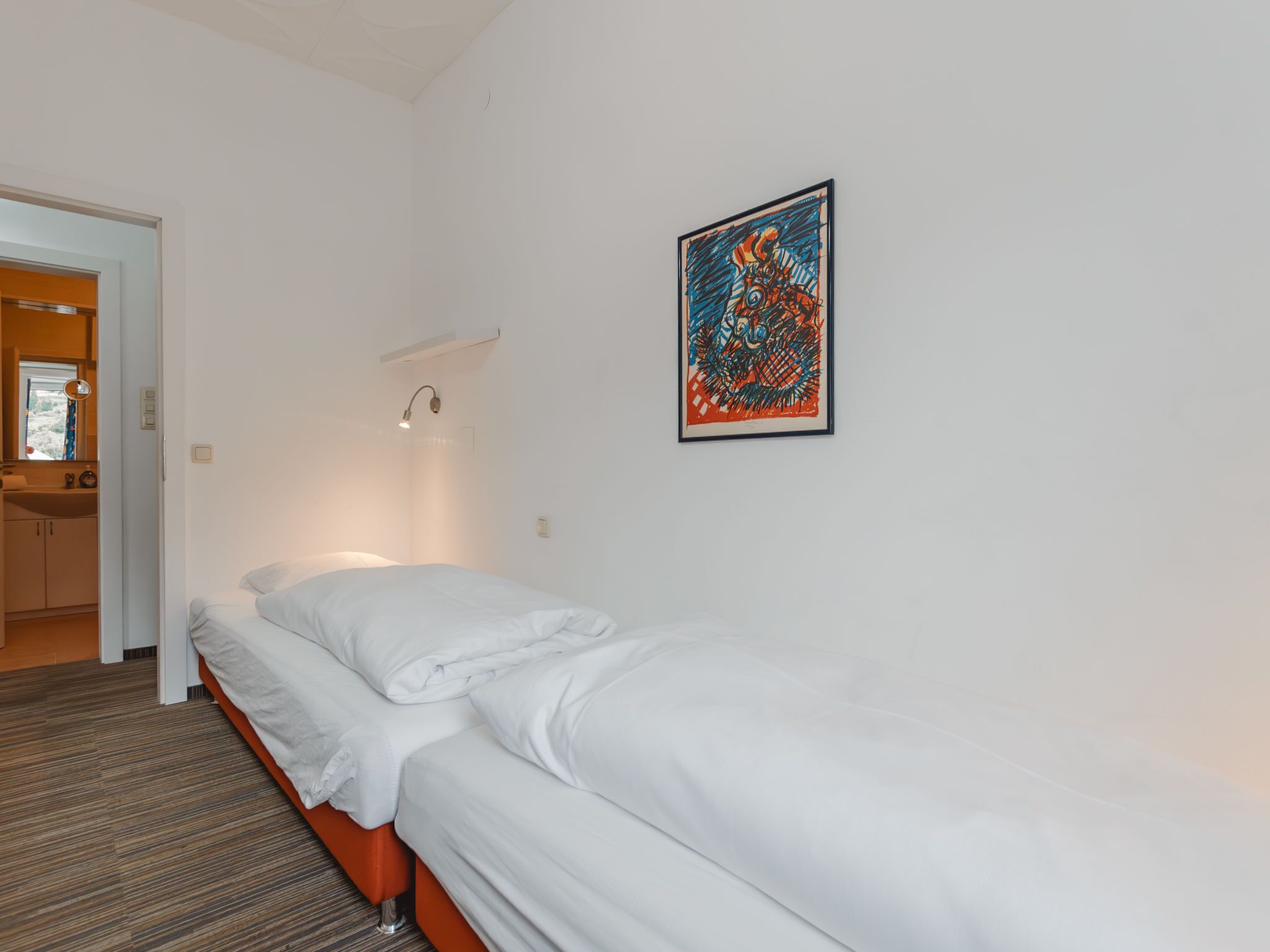 Een hotelkamer met een enkel bed, een klein nachtkastje, een wandlamp en een lijst met een abstract schilderij.