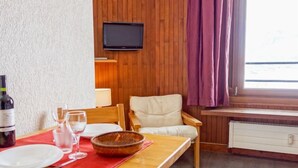 Een kamer met houten lambrisering, een eettafel gedekt voor twee personen, een aan de muur bevestigde televisie en een raam met paarse gordijnen.