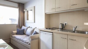 A compact hotel room with a sofa bed, a kitchenette with a sink and kettle, and a view of the city.