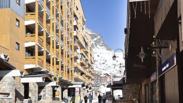 A multi-story building with balconies, a stone facade, and a mountainous backdrop.