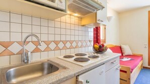 A compact kitchen with a sink, stove, and microwave. There's a small refrigerator and a bed with a red blanket in the adjacent room. - Chalet Club (Val Claret) by Interhome (Tignes)