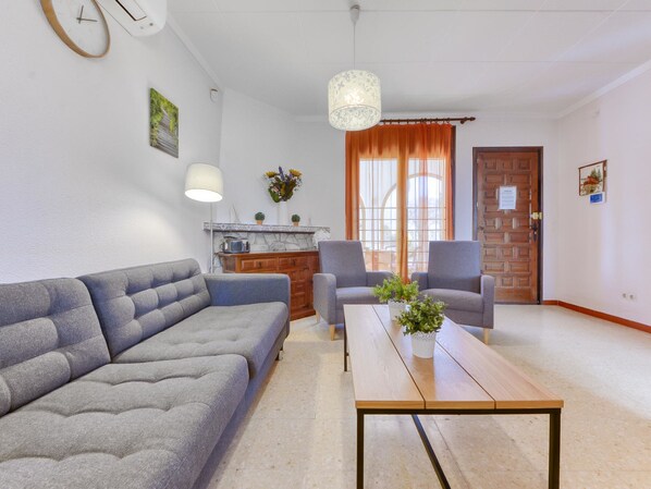 A modern living room with a grey sofa, two armchairs, a wooden coffee table, and a kitchen area with a stove and cabinets.