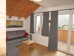 Un salon avec un canapé gris, une table basse en bois sur laquelle est posé un vase, et une vue sur un paysage montagneux, visible à travers une porte-fenêtre coulissante.