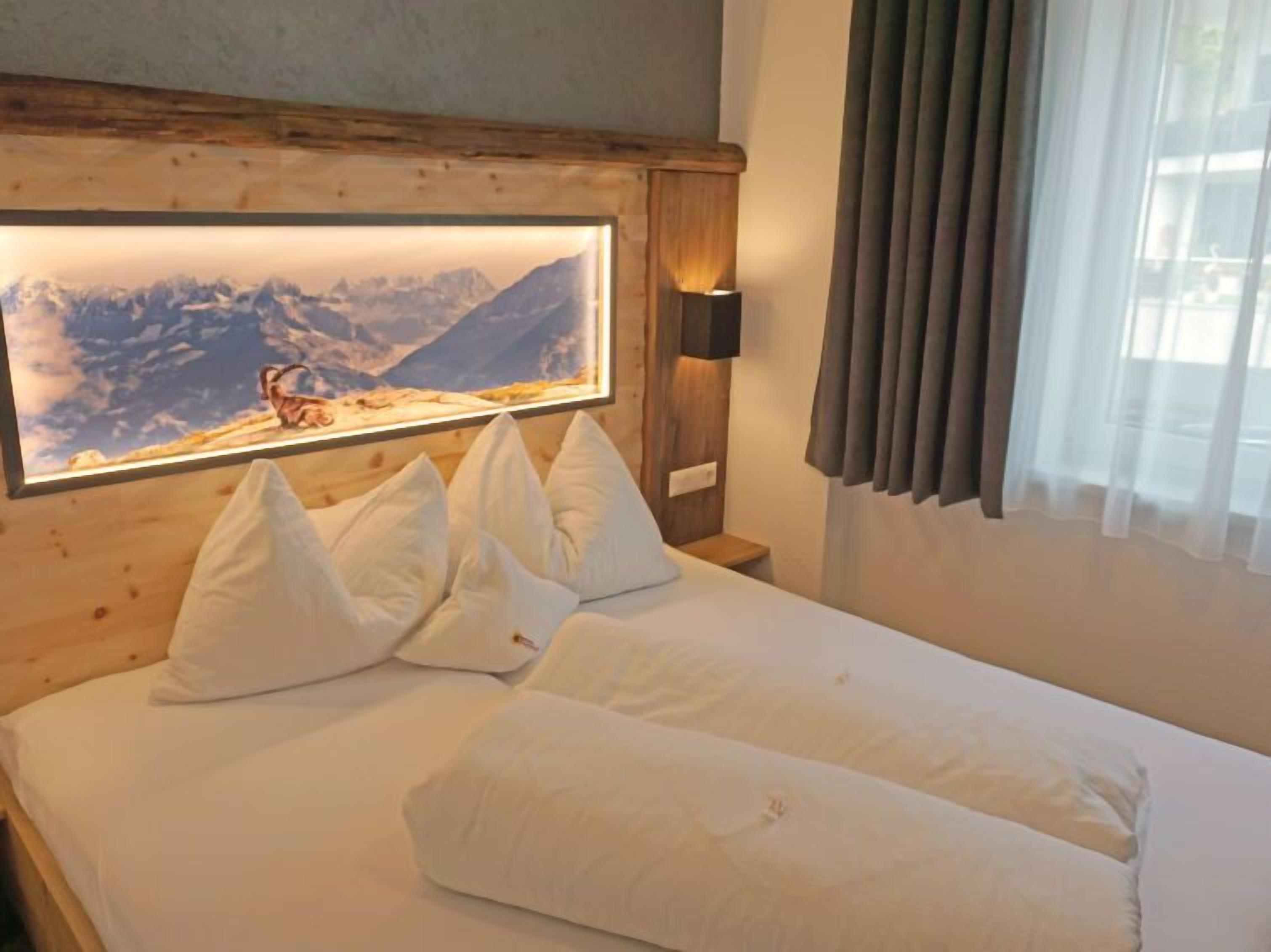 Ein Hotelzimmer mit Holz-Kopfteil, einem großen gerahmten Bild von einer Berglandschaft, weißer Bettwäsche und einer Nachttischlampe.