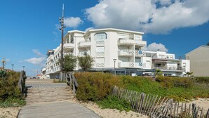 Un hotel frente al mar con varios balcones, un paseo marítimo de madera que conduce a la entrada y un cielo azul despejado.