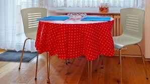Un comedor con una mesa redonda cubierta con un mantel rojo de lunares, dos sillas y una ventana con cortinas traslúcidas.