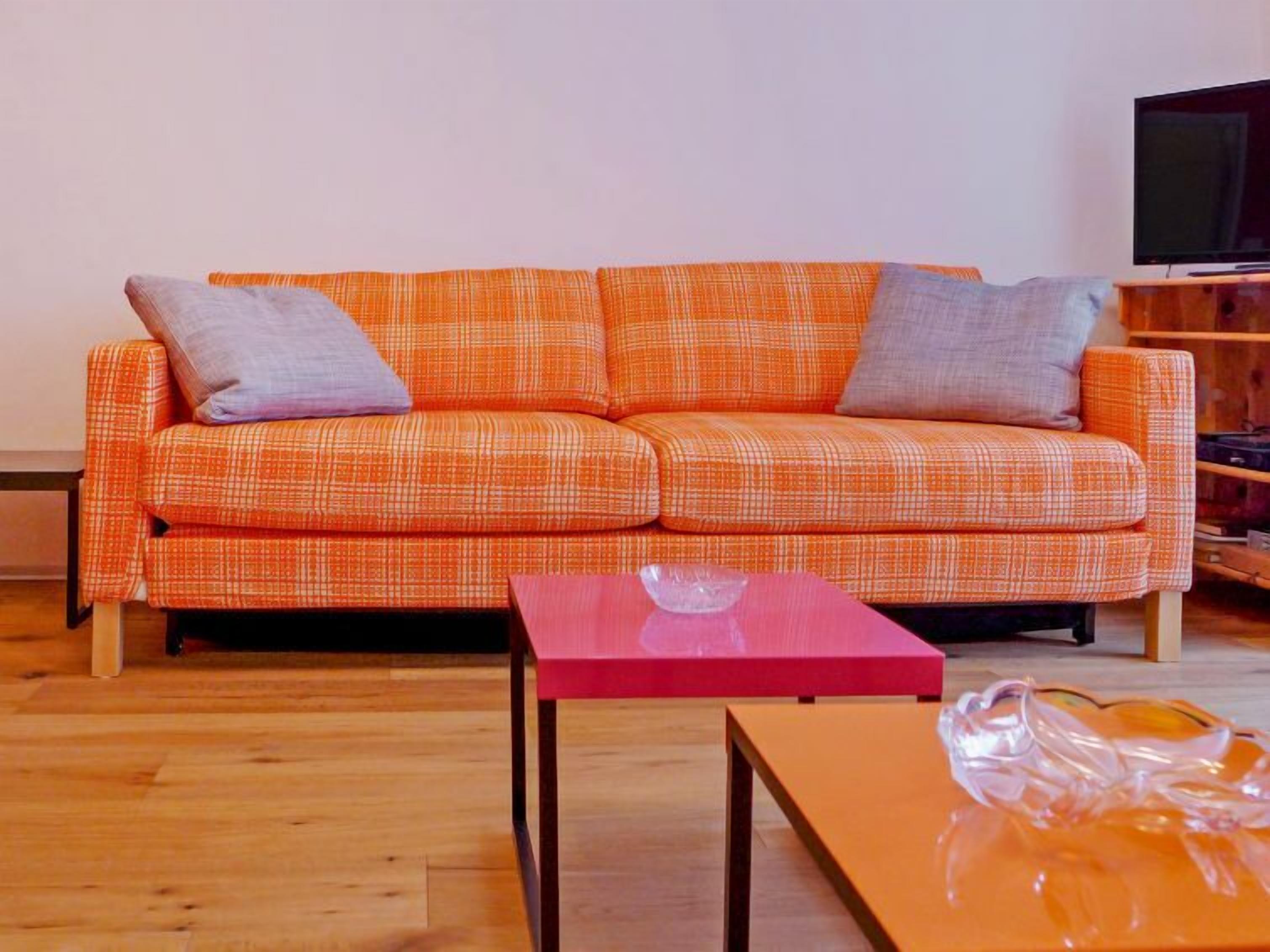 A living room with a plaid orange sofa, two grey cushions, a red coffee table, and a wooden floor.