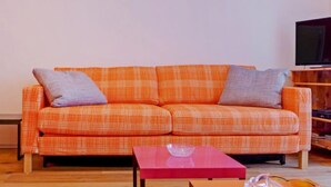 Una sala de estar con un sofá naranja estampado, dos cojines grises, una mesa de centro roja y piso de madera.