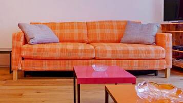 A living room with a plaid orange sofa, two grey cushions, a red coffee table, and a wooden floor.