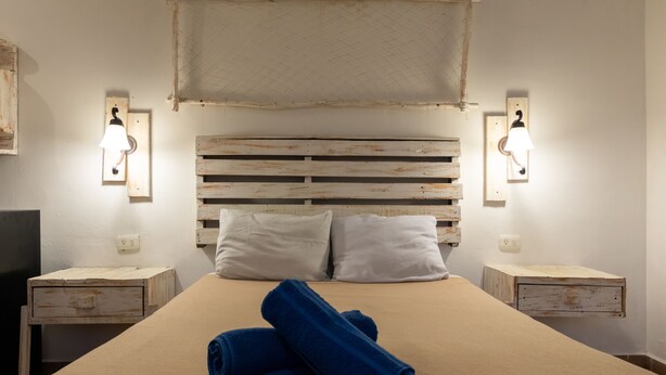 A bed with a wooden headboard, two white pillows, and a rolled blue towel on the bed. There are two bedside tables with light fixtures on either side of the bed. - Casa Isza (Playa del Carmen)