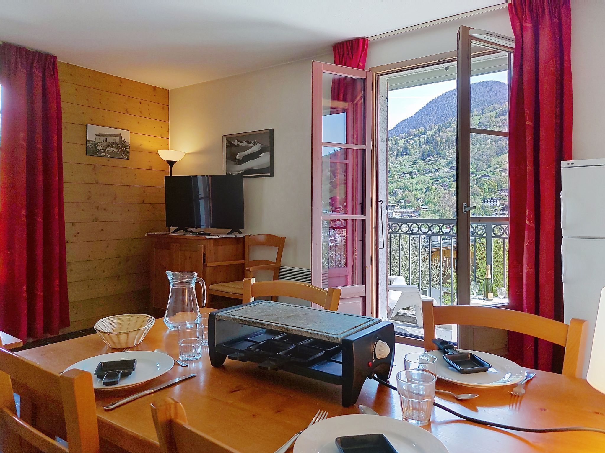 Zona pranzo con tavolo apparecchiato per due, televisione e vista su un paesaggio montuoso da un balcone.