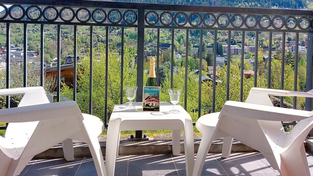 A balcony with a table and chairs, overlooking a mountainous landscape with a village below. - Les erables by Interhome (Saint Gervais)