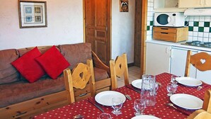 Een gezellige eethoek met een houten tafel gedekt voor vier personen, een bruine bank met rode kussens, en een keuken met witte kastjes en een magnetron.