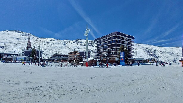 Een met sneeuw bedekte skiëresort met een meerlaags gebouw, skiliften en een helderblauwe lucht.