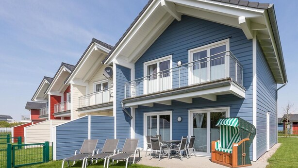 Een huis met twee verdiepingen, een balkon, een terras met stoelen en een tafel, en een groene omheining.