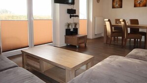 Een moderne woonkamer met een bank, een salontafel, een aan de muur bevestigde televisie, een eethoek met tafel en stoelen, en grote ramen met jaloezieën.
