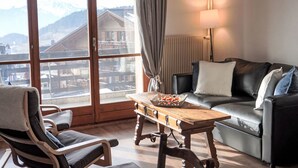 Una stanza con pavimento in legno, un divano in pelle, un tavolo in legno con un vassoio, una sedia e un'ampia finestra vista su edifici e montagne.