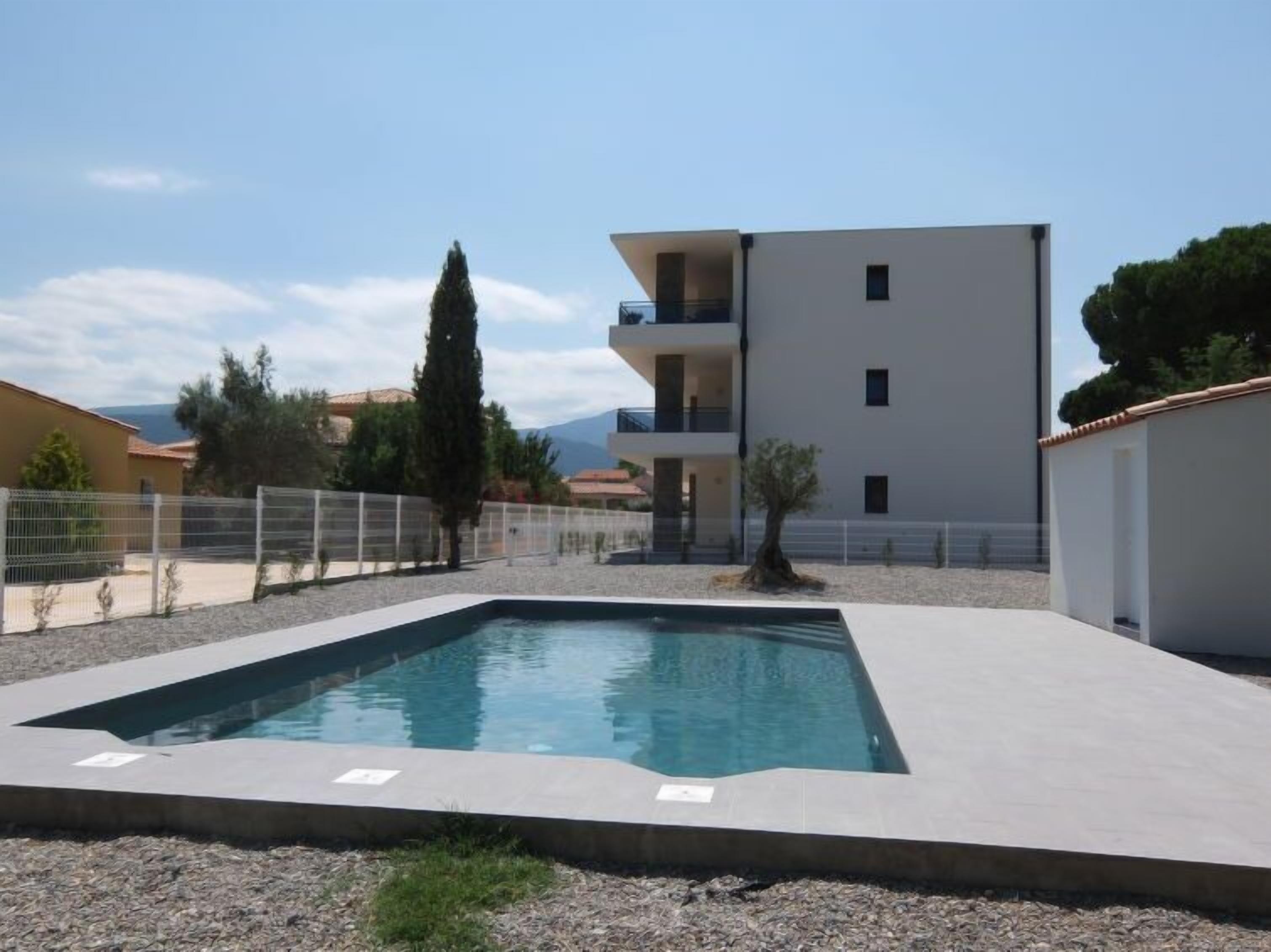 A modern building with a swimming pool, surrounded by a fence and vegetation.