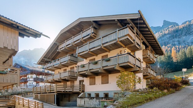Un immeuble à plusieurs étages, doté de balcons et d’une façade en pierre, avec une chaîne de montagnes en arrière-plan.