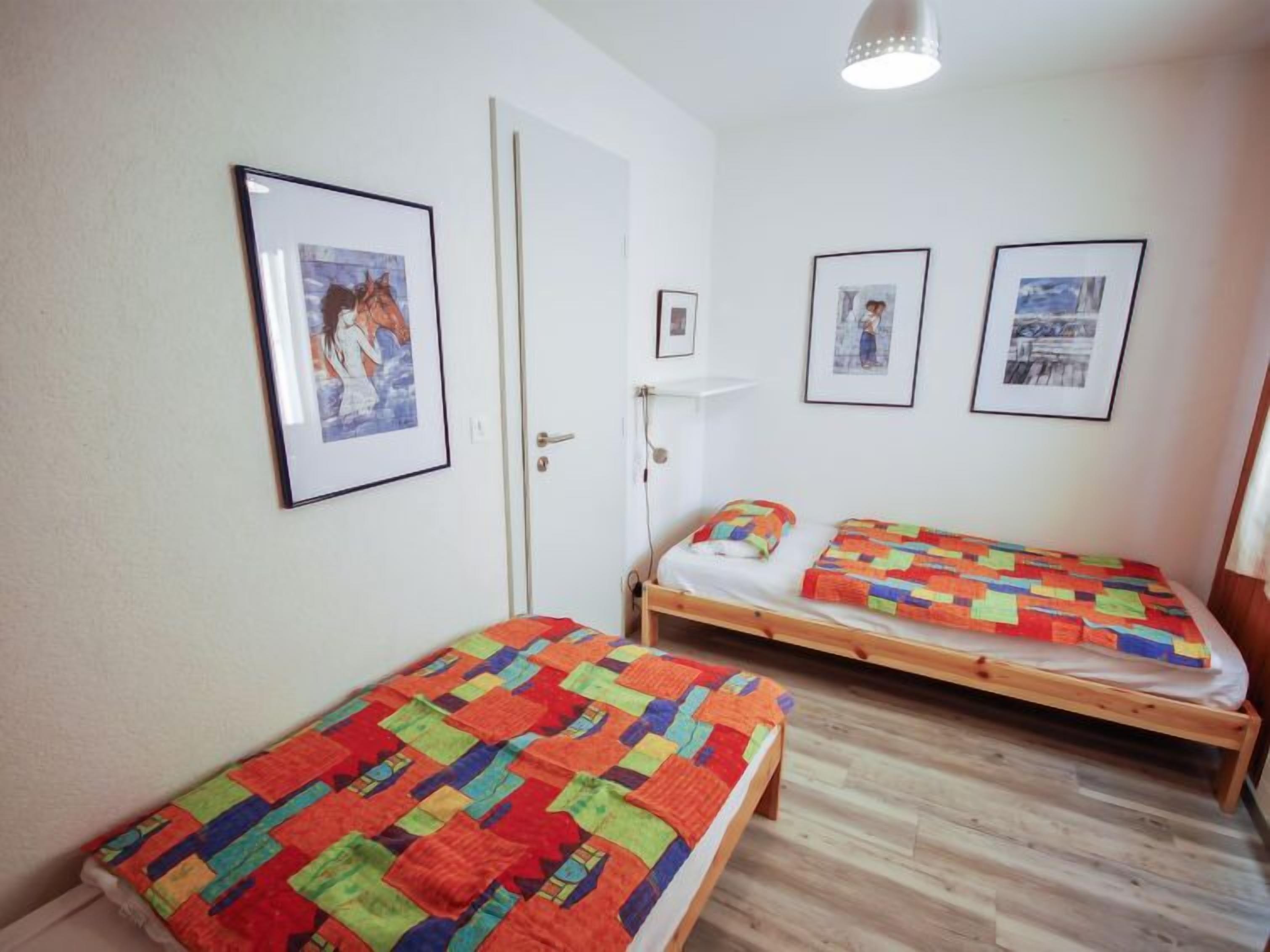 A room with two single beds, each with a colorful quilt. There are framed pictures on the walls, a white door, and a wooden floor.