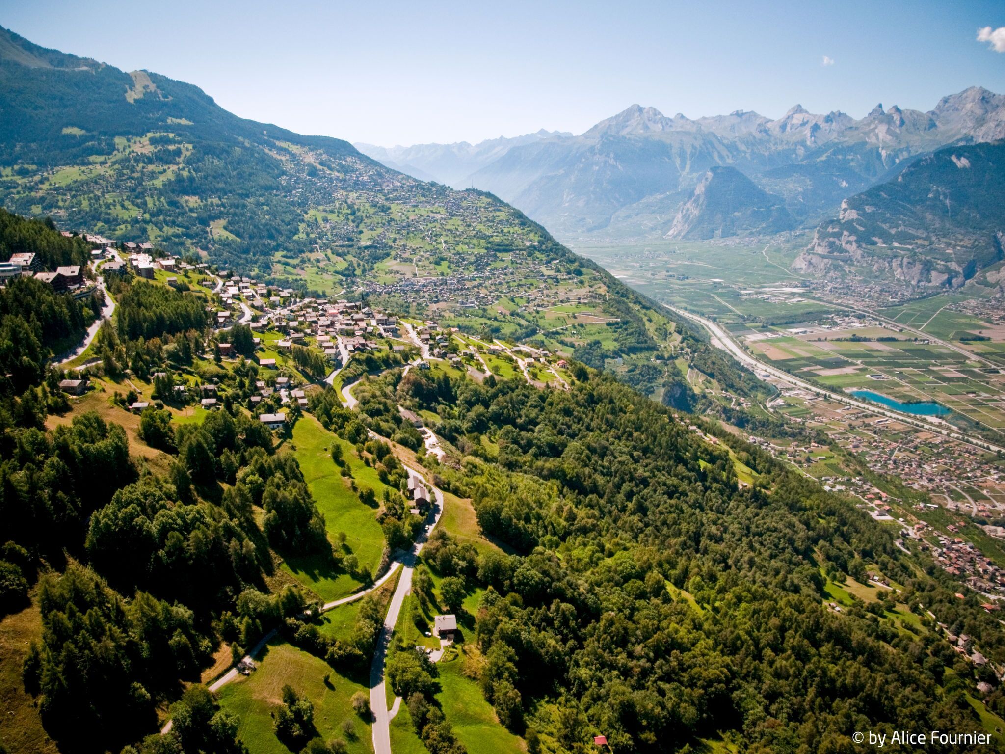 Vue aérienne d’un village de montagne, avec une route sinueuse, une végétation luxuriante et un petit plan d’eau.
